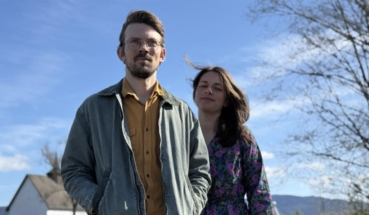 To personer smiler utendørs på en solfylt dag med klar blå himmel.