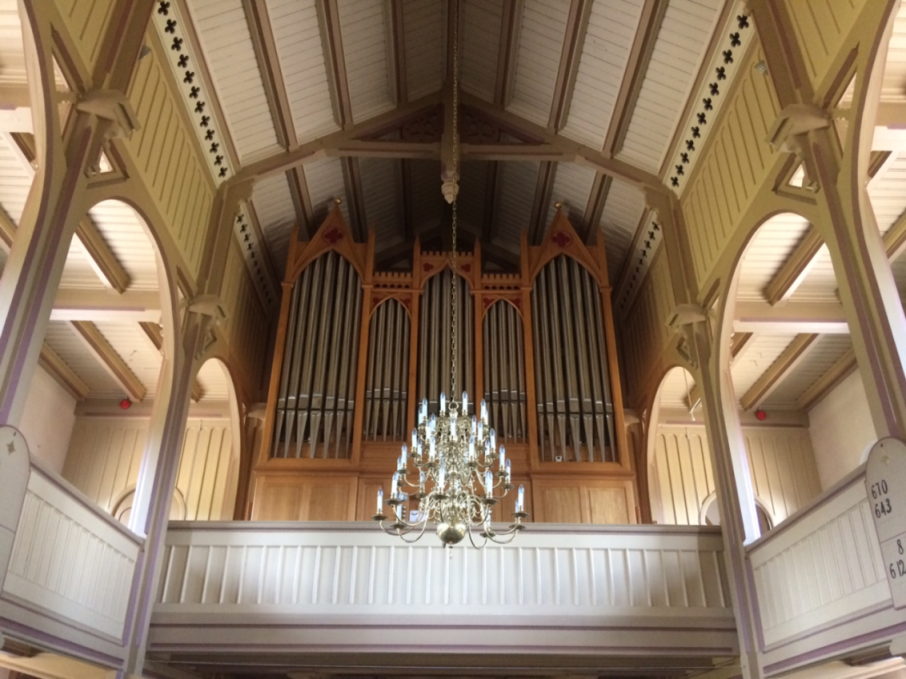Kirkeinteriør med pipeorgel og lysekrone, omgitt av trebuer og utskårne detaljer i taket.